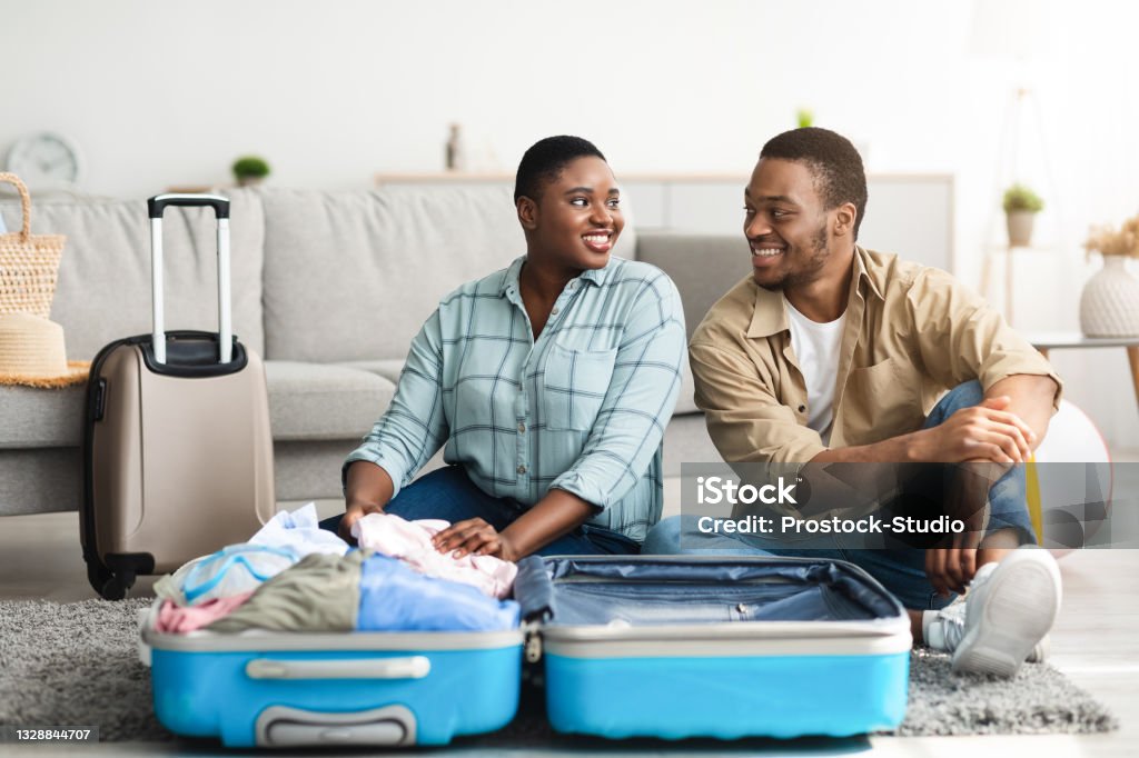 African American Couple Packing Suitcase Sitting On Floor At Home Family Vacation. African American Couple Packing Suitcase Smiling Each Other Sitting On Floor At Home. Happy Travelers Posing With Luggage Preparing For Journey. Tourism And Traveling Concept Suitcase Stock Photo