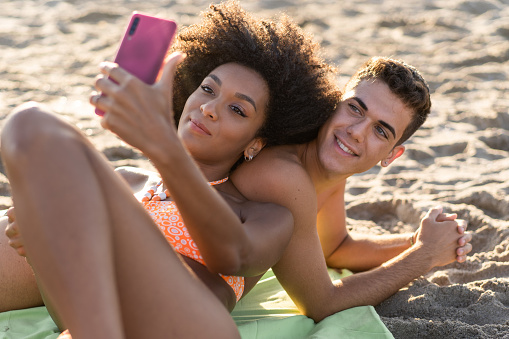 Generation z interracial couple lying down on the beach taking selfie with smartphone. Concept of young people from different races having fun on vacation using cellphones and new technologies