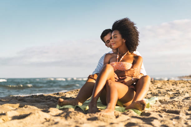 Interracial couple relaxing hugging together on a beach sitting on the sand stock photo
