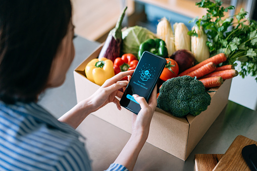Over the shoulder view of young Asian woman doing home delivery grocery shopping online with mobile app device on smartphone at home, with a box of colourful and fresh organic vegetables and fruits on the table