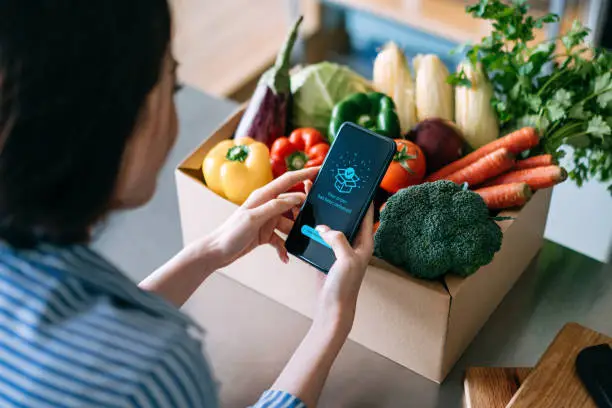 Photo of Over the shoulder view of young Asian woman doing home delivery grocery shopping online with mobile app device on smartphone at home, with a box of colourful and fresh organic vegetables and fruits on the table