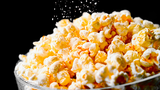 Close- up of popcorn in a glass bowl being sprinkled with salt against black background.