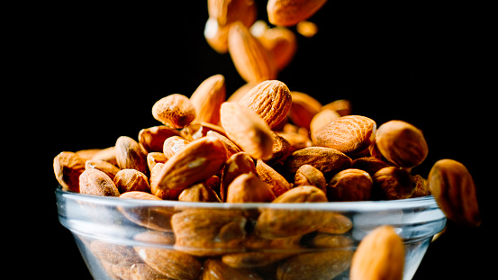 Close up of almonds falling into a glass bowl and overflowing against black background.