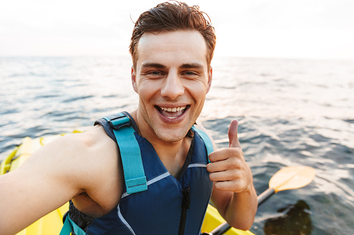 Image of young handsome man kayaking on lake sea in boat make selfie by camera showing thumbs up.