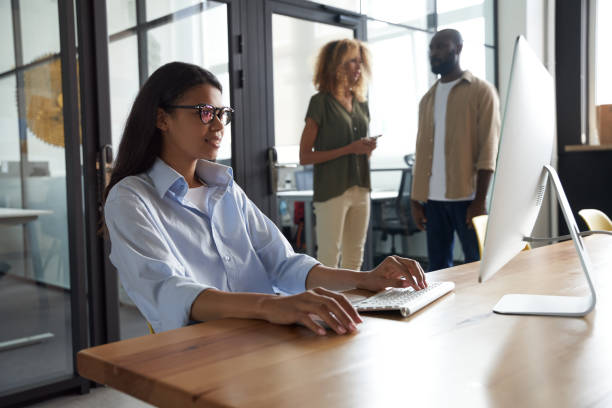 istock Young mixed race woman, female office worker working on computer while sitting at her workplace in the modern office, colleagues standing on the background 1328859095