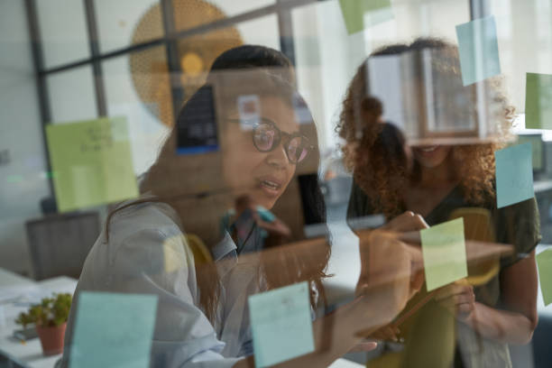 istock Brainstorming. Young mixed raced woman wearing glasses writing something on sticky notes on a glass board while working together with colleagues in the modern office 1328859107