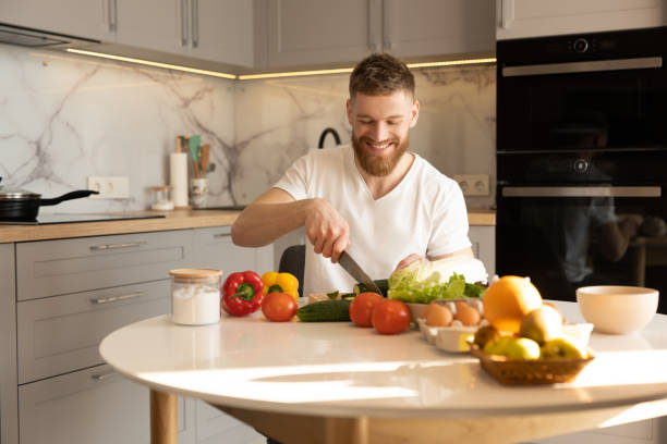 istock Young man sit at table and cut cucumber on salad 1328861834