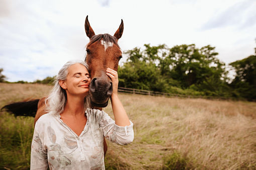 Beautiful mature woman enjoying with closed eyes her brown arabian mare in the free nature. Selective focus. Slightly grain. Part of a series.