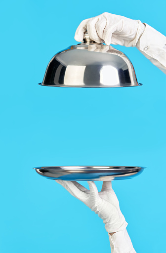 Elegant waiter's hands in white gloves holding silver tray and cloche on blue background. Restaurant, horeca, first class service concept