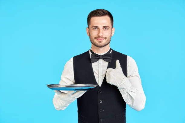 elegant young waiter holding empty silver tray and showing thumbs up sign over blue background - tray glove butler white imagens e fotografias de stock