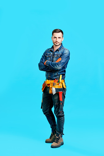 Full length portrait of confident handsome male construction worker staying over blue background looking to camera
