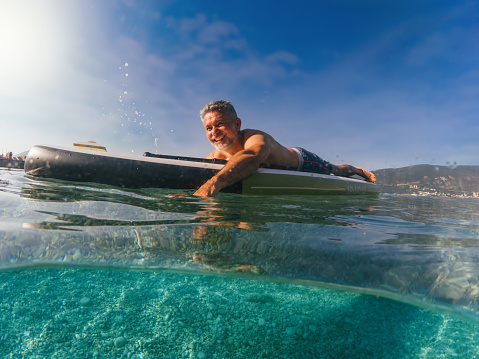 Joyful mature man is training on a SUP board on the beach on a sunny morning.