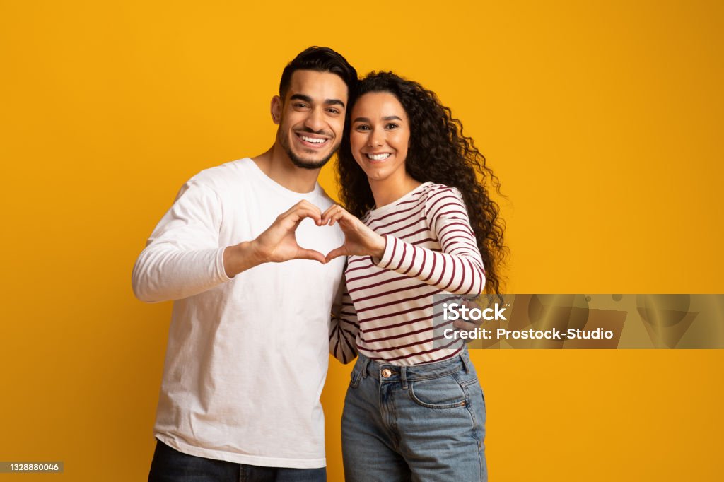 Portrait Of Romantic Arabic Couple Making Heart Gesture With Hands Together Portrait Of Romantic Arabic Couple Making Heart Gesture With Hands Together, Middle Eastern Man And Woman Connecting Their Fingers Into Love Sign, Smiling At Camera While Posing On Yellow Background Couple - Relationship Stock Photo