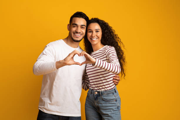 Portrait Of Romantic Arabic Couple Making Heart Gesture With Hands Together Portrait Of Romantic Arabic Couple Making Heart Gesture With Hands Together, Middle Eastern Man And Woman Connecting Their Fingers Into Love Sign, Smiling At Camera While Posing On Yellow Background middle eastern ethnicity photos stock pictures, royalty-free photos & images