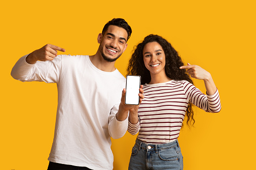 Happy Middle Eastern Couple Showing Blank Smartphone Screen For Mockup, Cheerful Arabic Man And Woman Demonstrating New Mobile App While Standing Together Over Yellow Background, Copy Space