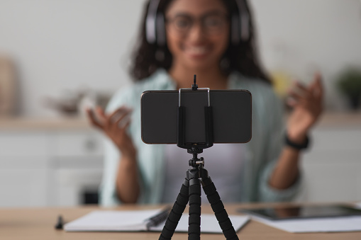 Devices for remote work at home, shooting video blog, new normal. Happy young african american lady in glasses and headphones gestures, talks and looks at webcam smartphone on tripod, selective focus