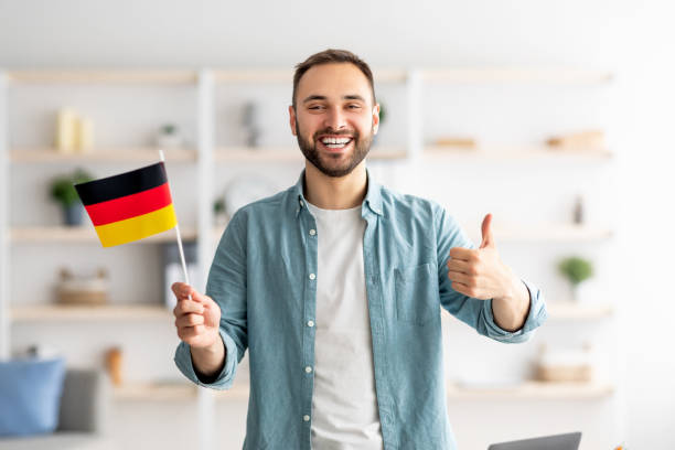 Hombre caucásico feliz mostrando el pulgar hacia arriba y la bandera de Alemania, posando y sonriendo a la cámara en el interior - foto de stock