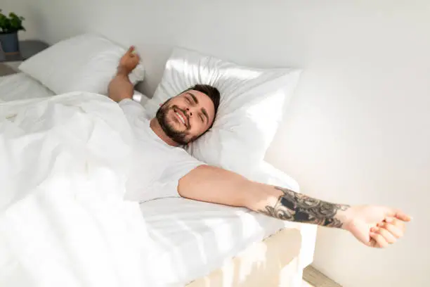 Comfortable sleep. Wellslept tattooed man waking up in morning and stretching hands, smiling with closed eyes while lying in bed, free space. Good morning concept