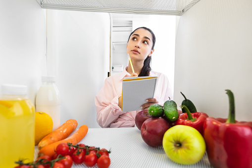 Portrait of young beautiful housewife looking at open refrigerator and thinking, holding notebook with pen taking notes making checklist of necessary food to buy or order, view from inside the fridge