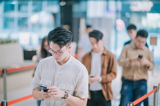 group of Asian Chinese young customer looking at smart phone while waiting in line in front ticket counter buying movie tickets and  popcorns drink contactless payment before movie show time at movie theater cinema