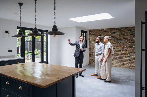 Wide angle view of Black couple listening to sales representative describing features of luxury home with exposed brick dining area and Shaker-style kitchen.