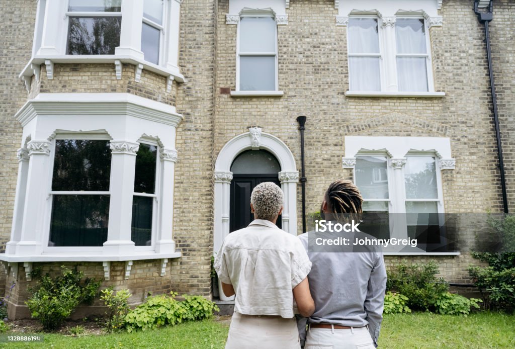Portrait of new homeowners admiring their investment Rear view of mid adult Black couple standing arm in arm and looking at facade of restored 1890 double fronted Victorian house. House Stock Photo