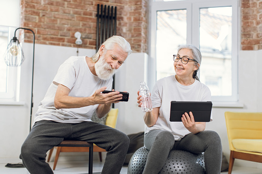 Smiling mature man and woman taking break during morning exercises and using modern tablet with smartphone. Concept of people, recreation and gadgets.