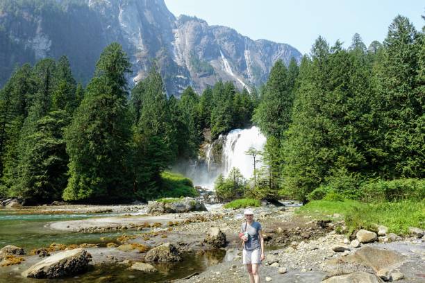 A woman standing on the shores with a waterfall, forests and a giant rock face behind her, on a beautiful summer day, in Princess Louisa Inlet, British Columbia, Canada. stock photo