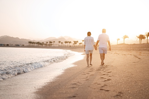 Happy mature senior couple walking and looking at each other on beach during sunset. Aging together and retirement lifestyle concept