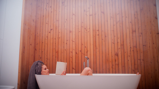 Modern, elegant bathroom with wooden wall and big bathtub. Woman reading and relaxing