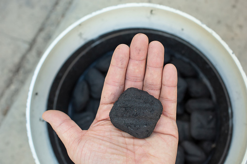 A top down view of a hand holding a single charcoal briquette over a bucket full of several briquettes.