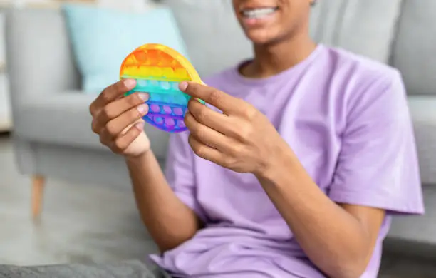 Photo of Unrecognizable black teen guy using POP IT toy to relieve stress at home, closeup