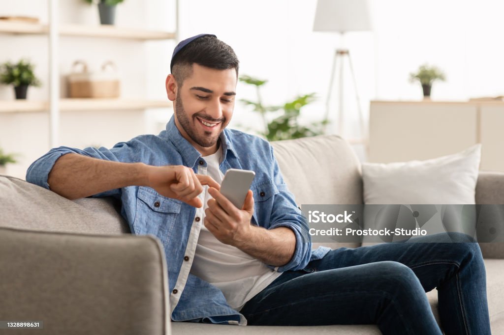 Smiling jew man using smart phone sitting on couch Useful App. Handsome Israeli Man In Kippah Using Smartphone While Relaxing On Couch At Home, Jewish Guy Sitting On Sofa In Living Room, Browsing Internet Or Messaging With Friends, Free Copy Space Mobile Phone Stock Photo