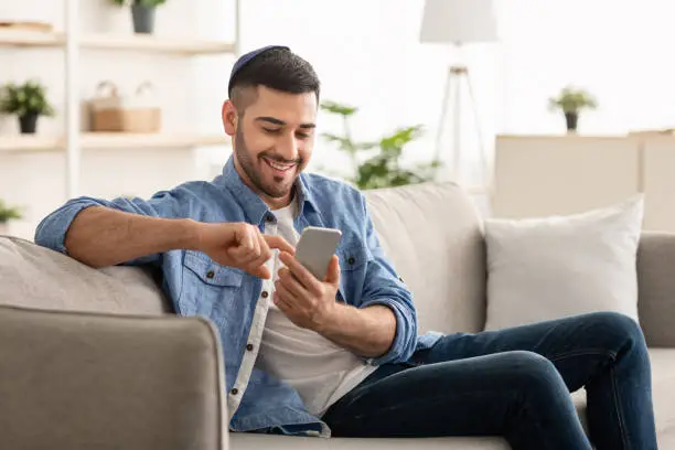 Useful App. Handsome Israeli Man In Kippah Using Smartphone While Relaxing On Couch At Home, Jewish Guy Sitting On Sofa In Living Room, Browsing Internet Or Messaging With Friends, Free Copy Space