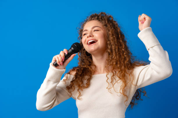 istock Young joyful curly-haired woman singing in microphone against blue background 1328899137