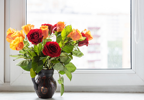 fresh red and yellow roses in ceramic jug on window sill at home with cityscape on background