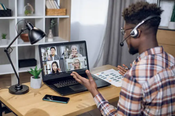 Multiethnic group of people having business meeting through video call on laptop. View from shoulder of black man sitting at home office and working remotely.