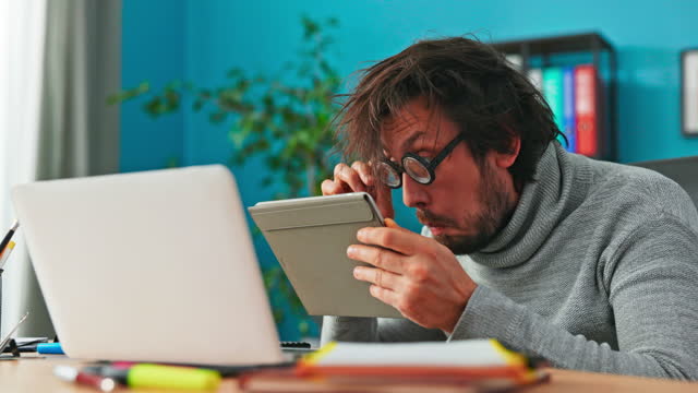 Curious, strange man working in office looks into a tablet, brings glasses closer to screen to see better, has puzzled look on face, shocked, surprised, looks at camera with delight while shining eyes
