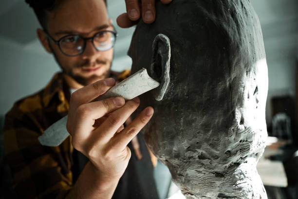 istock Young man sculpting head out of clay in studio working on ear with tool 1328918213