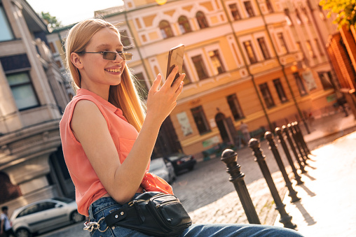 Young cheerful blonde girl smiles and uses the phone while walking around the city