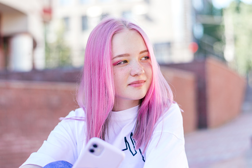 La adolescente hipster de pelo rosado positivo con una camiseta blanca está usando un teléfono inteligente en un día de verano en la calle de la ciudad. Concepto de verano. Estilo generación Z. Concepto de redes sociales photo