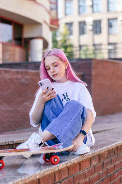 A pink-haired hipster teenage girl in a white T-shirt,jeans and with a skateboard is sitting on a city street on a summer day and using a smartphone.Generation Z style,social network,hobby. una adolescente hipster de pelo rosa con una camiseta blanca, pantalones vaqueros y con un monopatín está sentada en una calle de la ciudad en un día de verano y usando un teléfono inteligente. estilo generación z, red social, pasatiempo