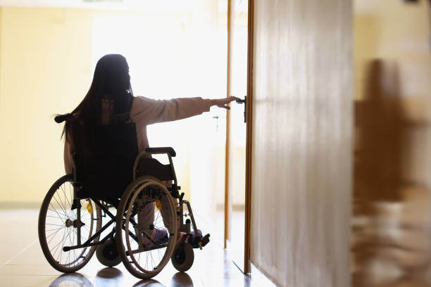 donna in sedia a rotelle si allunga per aprire la maniglia della porta nel corridoio buio - disablement foto e immagini stock