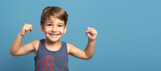 vista frontal del pequeño niño caucásico de cuatro años de edad de pie frente a la toma de estudio de fondo azul de pie confiado flexionando los músculos sonrientes creciendo y la fuerza y el concepto de salud - 4 5 years fotografías e imágenes de stock