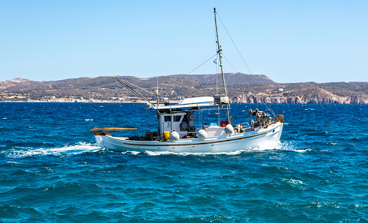 Cyclades, Greece. Fishing boat white traditional wooden vessel sailing in rough wavy sea. Rocky land, Greek island coast and clear blue sky background.