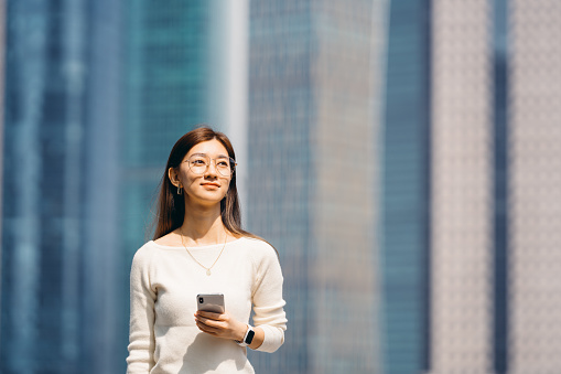 Confidence and professional young Asian businesswoman holding smartphone while standing against high rise corporate buildings in financial district in the city.