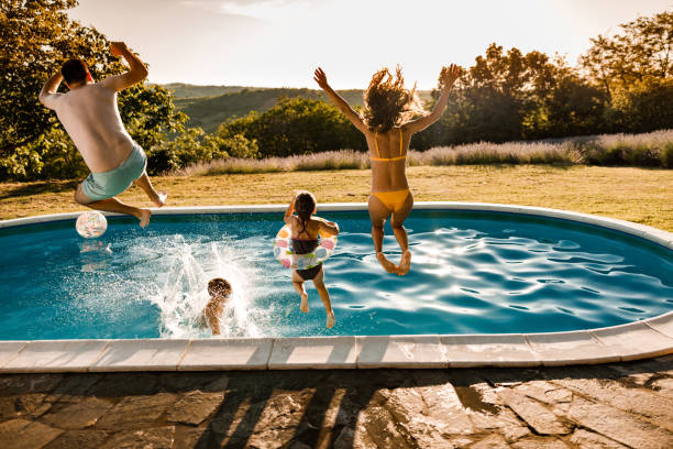 Back view of carefree family jumping in the pool at the backyard. Rear view of playful parents and their kids having fun while jumping into the pool during summer day at the backyard. Copy space. back yard stock pictures, royalty-free photos & images