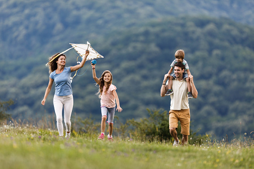 Playful family having fun while running in nature. Copy space. Father is carrying boy on shoulders while girl is holding a kite.
