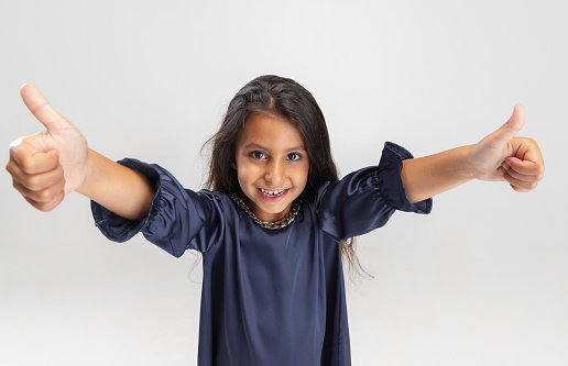 Thumbs up. One cute smiling preschool girl in beautiful blue dress posing isolated over white studio background. Childhood, fun, emotions, facial expression concept.