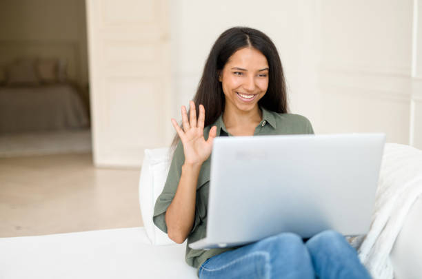 istock Young cheerful multiethnic woman waving hello to online interlocutor, using laptop 1328998899